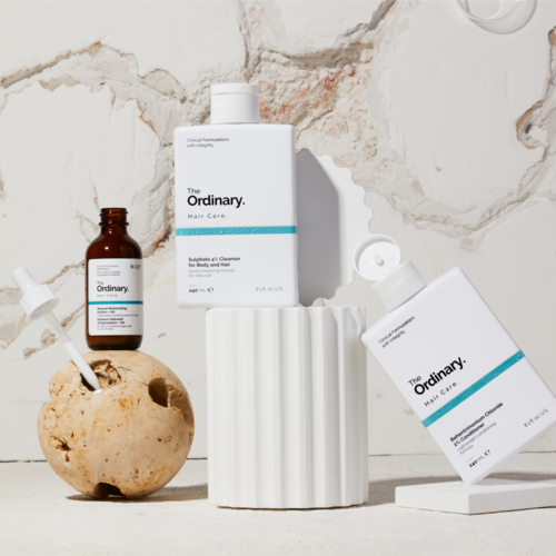The image shows skincare bottles and a brush in a minimalist setting with a textured white background.