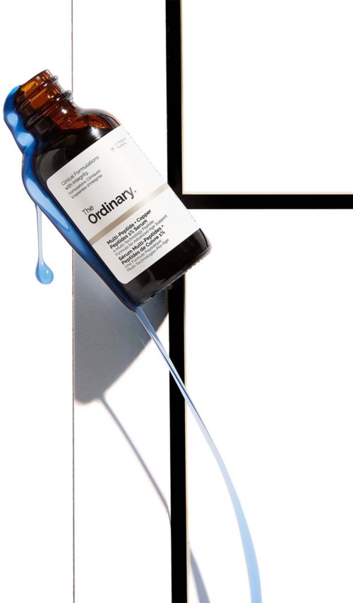 Een bruin glazen flesje met label 'The Ordinary', hangt schuin op een witte deur, met een blauwe druppel vloeistof die eruit valt.