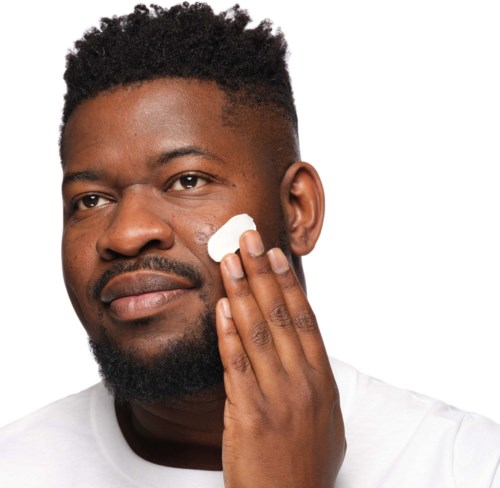 Een man brengt lotion aan op zijn gezicht met zijn hand, ziet er kalm en tevreden uit. Hij heeft kort krullend haar en een baard.