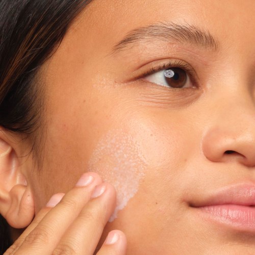 A woman applies a white cream to her cheek, smiling gently. Her skin looks smooth and clear.