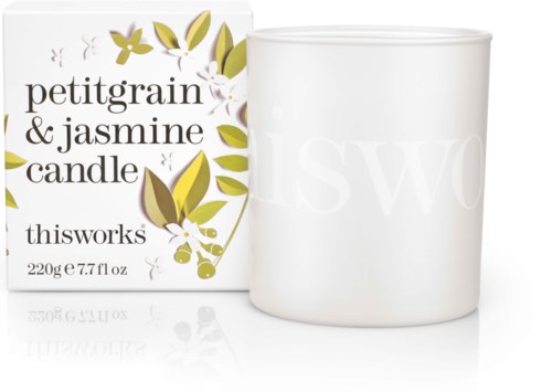 Een witte kaars in een pot naast een doos met 'petitgrain & jasmine candle' en groene en gele bladeren.