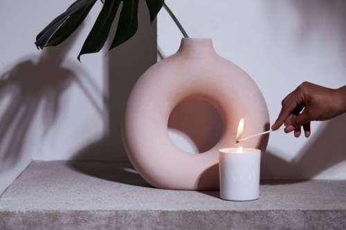 Iemand steekt een kaars aan naast een witte decoratieve vaas met een grote opening, op een grijze plek met een plant op de achtergrond.