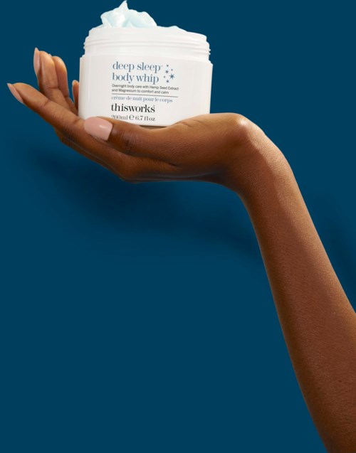 Een hand houdt een witte pot huidverzorgingscrème vast tegen een blauwe achtergrond. Het label zegt "deep sleep body whip."