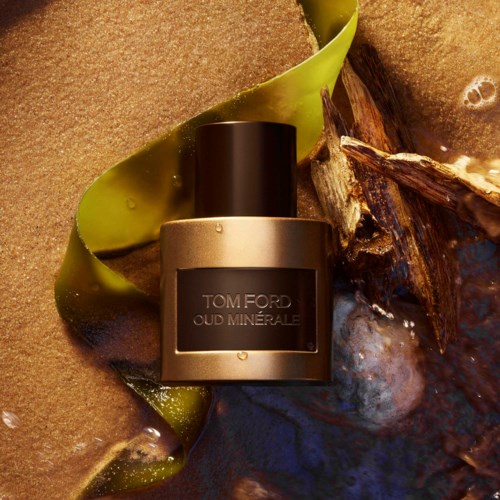 Een Tom Ford Oud Minerale parfumfles, omgeven door zand, zeewier en drijfhout op het strand.