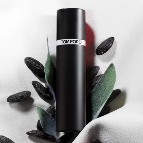 A black cylindrical bottle with a white band labeled 'TOM FORD' is surrounded by black seeds and green leaves on a white background.