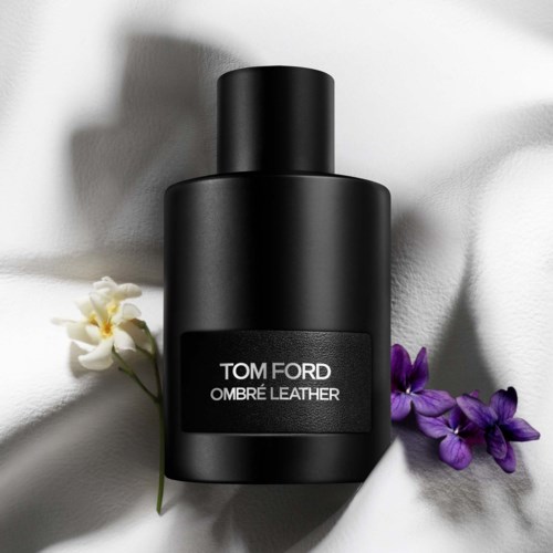 Eine schwarze Flasche von Tom Ford Ombré Leather-Parfüm auf weißem Stoff, umgeben von violetten Blumen.