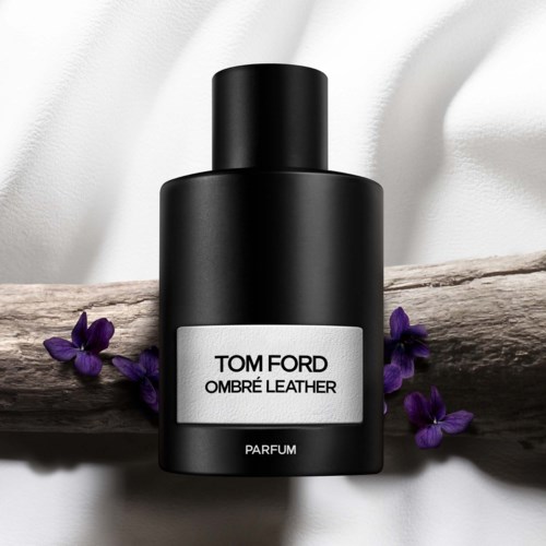 Zwarte fragrancefles met label 'Tom Ford Ombré Leather' en paarse bloemen eromheen op een houten stam.