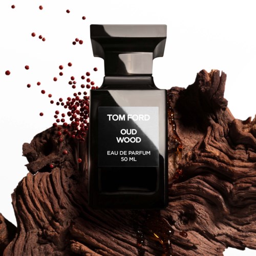 A black perfume bottle labeled 'Tom Ford Oud Wood' sits on a wooden surface with small red particles around it.