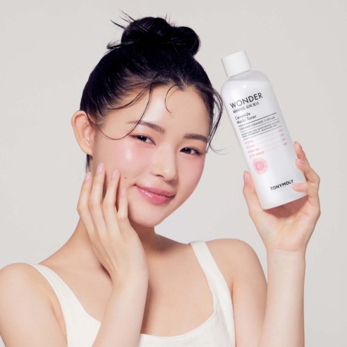 En kvinna med uppsatt hår som ler och håller en hudvårdsflaska märkt 'Wonder' av Tony Moly.