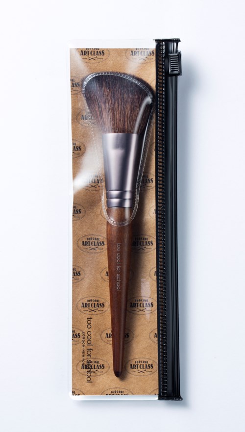 Ein Make-up-Pinsel mit braunem Griff in Verpackung neben einem schwarzen Lineal. Die Verpackung hat Texte und Muster.