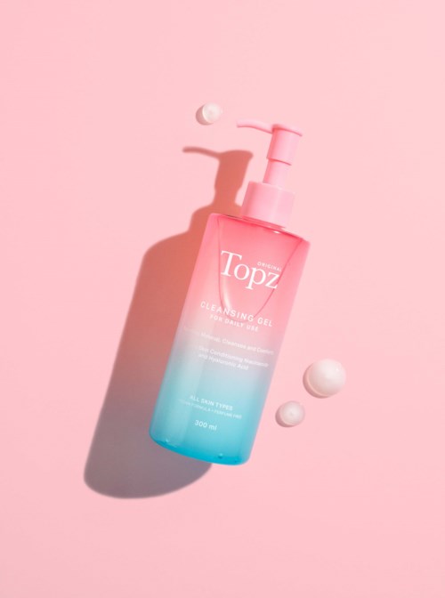 A pink and blue bottle of Topz cleansing gel, with little foam balls nearby, on a pink background.