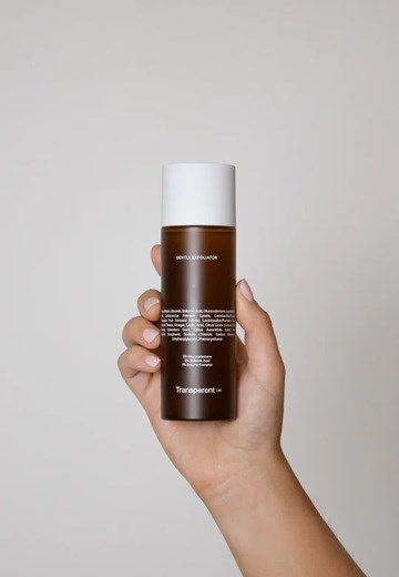 Eine Hand hält eine braune Hautpflegeflasche mit weißer Kappe, Text auf dem Etikett und einfacher grauer Hintergrund.