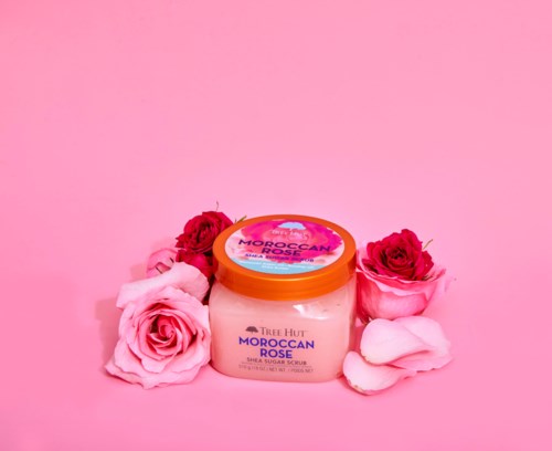 Ein Glas Tree Hut Moroccan Rose Shea Scrub umgeben von rosa und roten Rosen auf rosa Hintergrund.