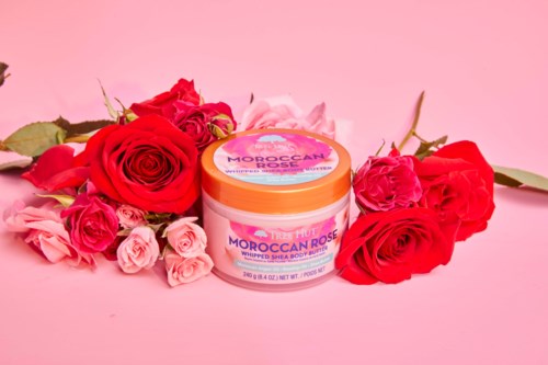 Ein Glas Moroccan rose whipped shea body butter umgeben von rosa und roten Rosen auf rosa Hintergrund.