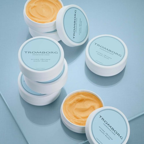 Acht Behälter mit gelblich wirkendem Balsam oder Creme, beschriftet 'TROMBORG' mit blauen Deckeln, auf blauer Oberfläche.