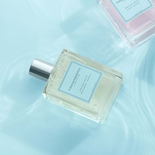 Zwei Parfümflaschen liegen auf einer hellblauen Oberfläche, reflektieren sanft. Eine ist transparent, die andere ist pink. Beide haben Sprühköpfe.