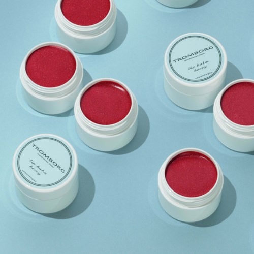 Kleine runde Behälter mit rotem Lippenbalsam, einige mit weißen Etiketten, andere ohne, auf hellblauer Oberfläche.