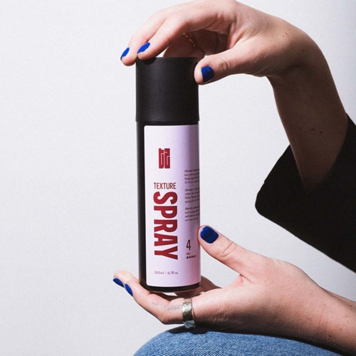 En persons händer håller en svart och vit sprayburk med etiketten 'Texture Spray'.
