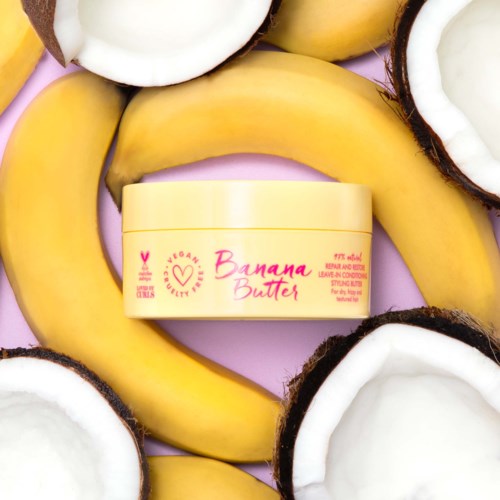 Ein Behälter Bananenbutter umgeben von Bananen und Kokosnüssen auf rosa Hintergrund.