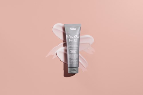 A grey tube of skincare product with white text is placed on a pink background with soft white flowers.