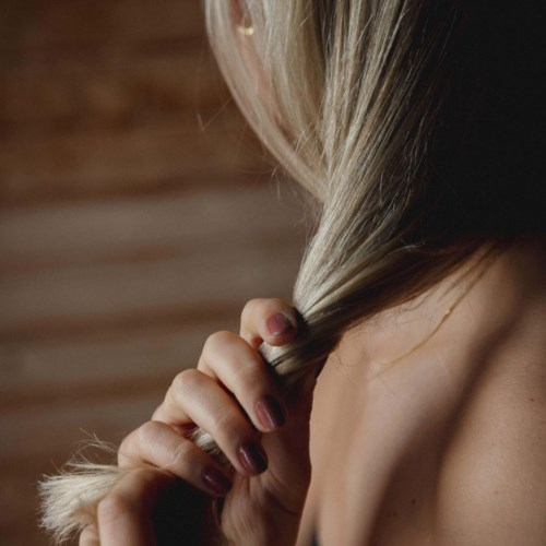 Iemand houdt een blond haar in de hand, een deel van de schouder zichtbaar, tegen een vage bruine achtergrond.