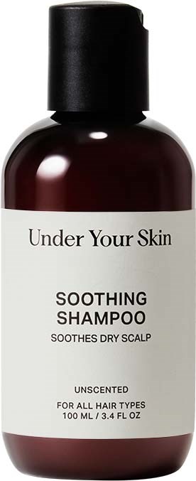 Een donkerbruine fles met wit etiket en zwarte dop, gelabeld "Under Your Skin Soothing Shampoo" voor een droge hoofdhuid.
