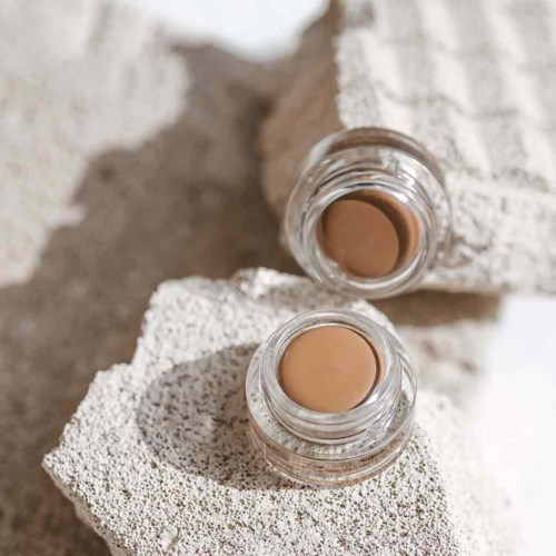 Zwei kleine Gläser mit beiger Creme auf strukturierten, weißen Steinen, mit einem Handtuch im Hintergrund.