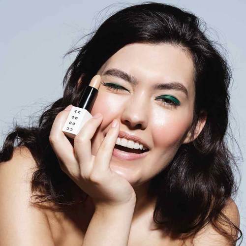 Eine Frau mit dunklen Haaren, die Make-up macht, lächelt und Lippenstift hält.