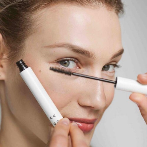 Eine Frau trägt Mascara auf ihre Wimpern auf, hält eine Mascara und eine Bürste.