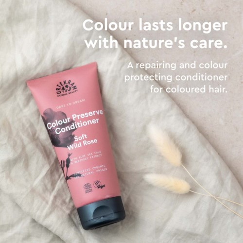 Pink bottle of Colour Preserve Conditioner with wild rose. Text about caring for colored hair. Soft fabric background with decorative feathers.