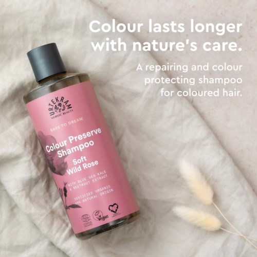A shampoo bottle with pink label and black cap, placed on white fabric with decorative plants. Text highlights benefits and ingredients.