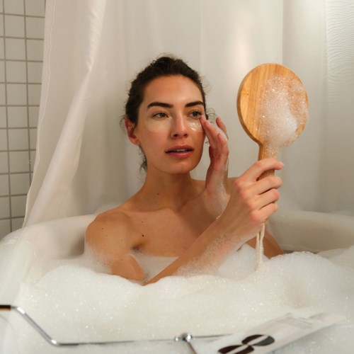 Eine Frau mit dunklem Haar und klarer Haut badet, hält eine Holzbürste, umgeben von Seifenblasen.