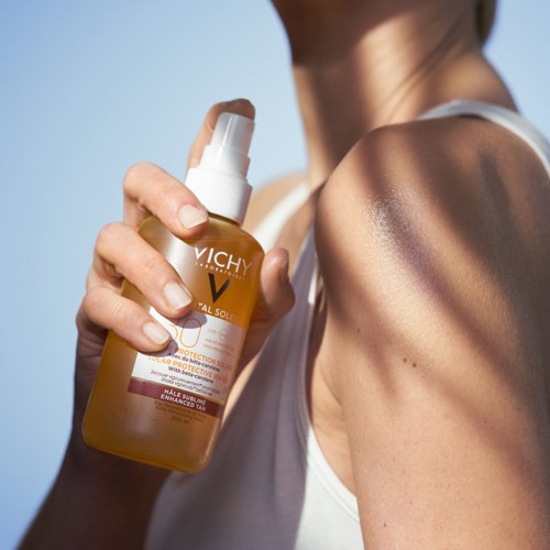 A woman sprays sunscreen on her shoulder outdoors on a sunny day.