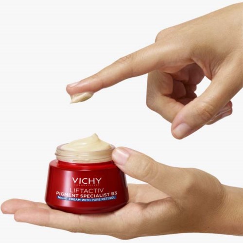 Eine Hand hält einen roten Vichy-Topf mit Creme, die zum Gebrauch entnommen wird.