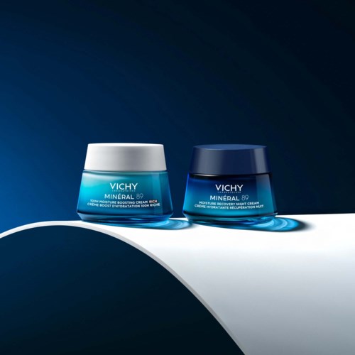 Two jars of Vichy skincare cream are displayed on a curved white surface with blue and dark background.