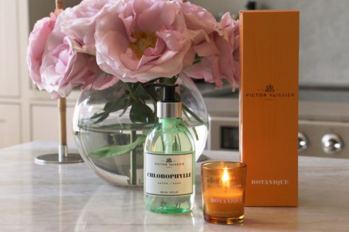 Ein Parfümflasche, eine brennende Kerze, eine Schachtel und rosa Blumen in einer Glasvase auf einer Küchenarbeitsplatte.