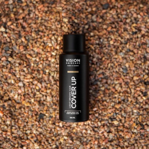 Ein schwarzer Behälter mit der Beschriftung 'VISION HAIRCARE' und 'COVER UP' auf kleinen, bunten Steinen.