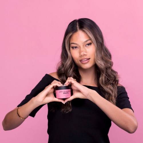 Eine Frau mit langen, welligen Haaren hält ein pinkes Glas mit der Aufschrift "dry shampoo paste" vor einem pinken Hintergrund.