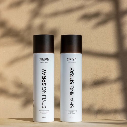 Two cylindrical bottles with black caps labeled 'Styling Spray' and 'Shaping Spray' stand on a surface. Shadows are cast in sunlight.