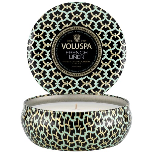 En rund ljusbehållare med ett svart och guld invecklat mönster, etiketten säger 'Voluspa French Linen'.