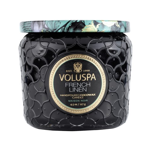 Svart burk med blomsterlock som innehåller ett doftljus, märkt som Voluspa French Linen, handgjort av kokosvax.