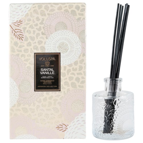 Eine dekorative Schachtel und ein Glastopf mit schwarzen Stäbchen für Duft. Die Box trägt die Aufschrift Voluspa Santal Vanille.