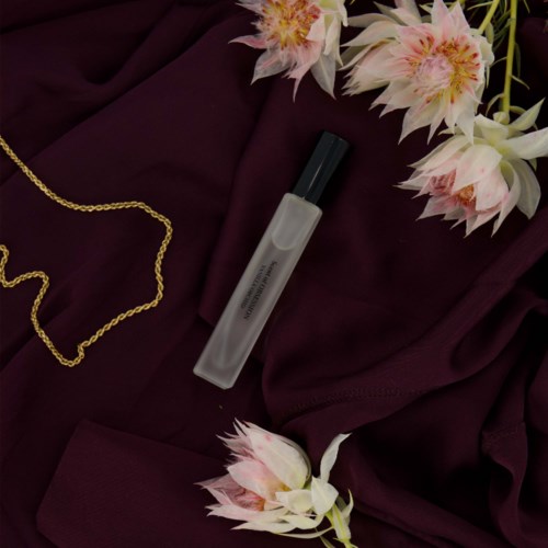 A perfume spray bottle on dark fabric surrounded by pink and white flowers and a gold chain.
