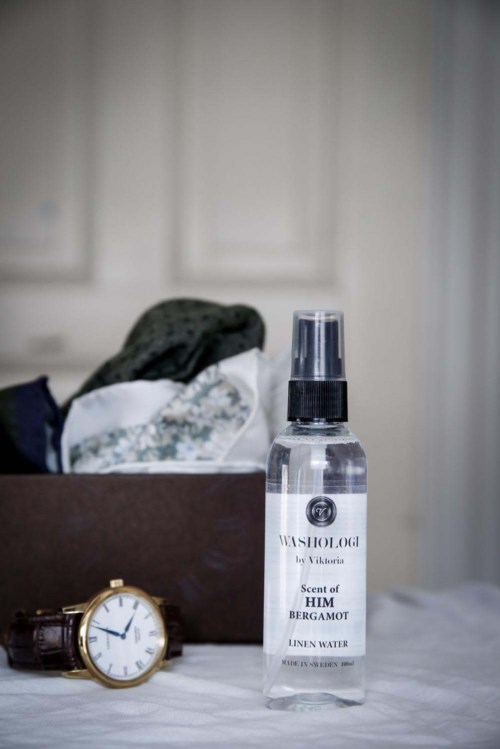 A bottle of linen water scent with bergamot sits on a table, with a gold watch and a box of clothes behind.