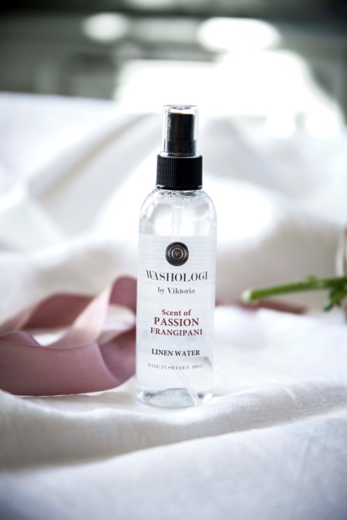 A clear bottle of linen water spray labeled 'Scent of Passion Frangipani' with a black spray top, on white fabric.