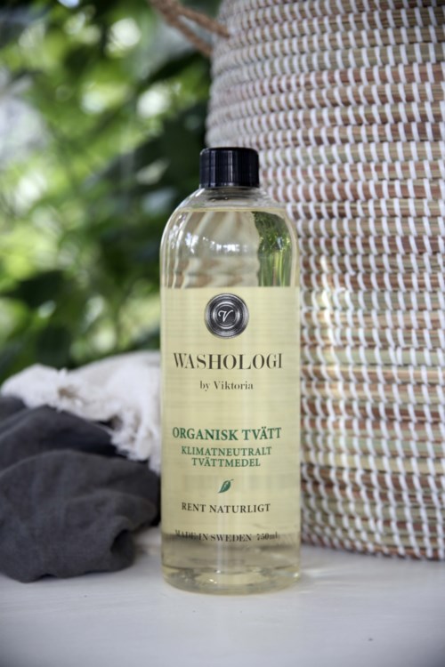 A clear bottle labeled Washologi with eco-friendly laundry detergent on a surface, with cloth and a woven basket nearby.