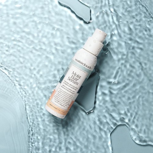Een witte fles met 'Moist Hair Serum' ligt op een gestructureerd, ijskoud blauwe oppervlak met ijsblokjes eromheen.