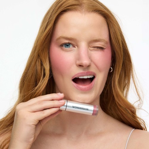 A woman with red hair holds a Wonderskin lip gloss tube, winking and smiling against a white background.