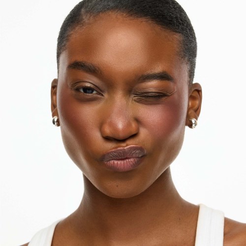 A woman with short hair and earrings is winking and pursing her lips against a white background.