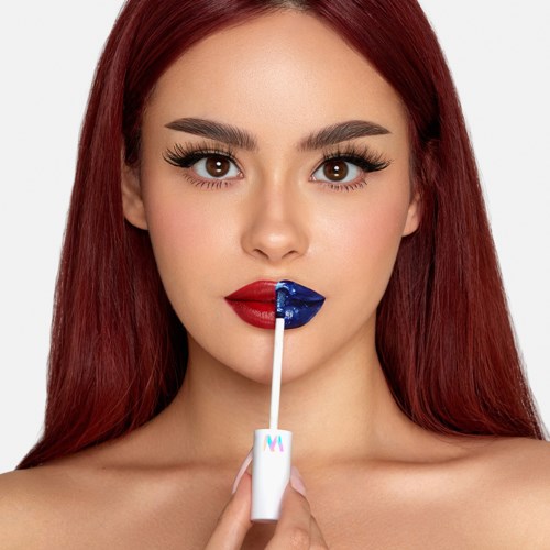 Eine Frau mit roten Haaren trägt dunkle blaue Lippenstift mit einem Applikator auf. Sie schaut direkt in die Kamera.